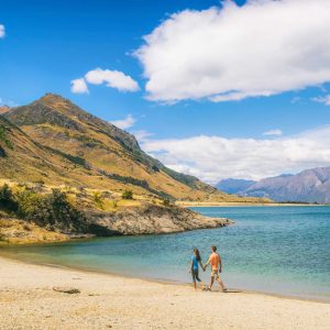 New Zealand travel people tourists visiting Otago region walking on shore of lake Hawea nature landscape. Man and woman happy at beach Near Wanaka. Young tramping hikers adventure lifestyle.